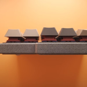 May include: A mechanical keyboard side profile showing a textured gray frame and a row of keys with a mix of light beige and dark gray keycaps. The switches beneath the keycaps are a deep red color, visible between the frame and the keycaps.