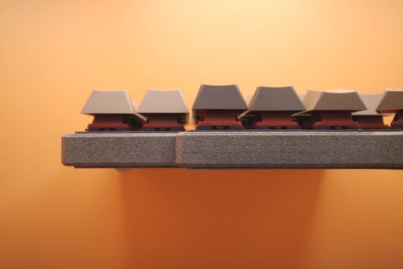 May include: A mechanical keyboard side profile showing a textured gray frame and a row of keys with a mix of light beige and dark gray keycaps. The switches beneath the keycaps are a deep red color, visible between the frame and the keycaps.