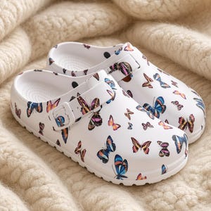 Puede incluir: Un par de zuecos blancos sin cordones con un estampado de mariposas de colores. Cada zapato incluye una tira ajustable funcional en la parte superior con una hebilla blanca a juego, punta redondeada y una plantilla texturizada.