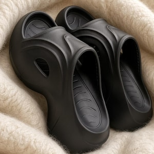 May include: A pair of black slip-on sandals featuring a thick, molded platform sole and a smooth, matte finish. The upper design includes curved, raised ridges and side cutouts, with a contoured footbed displaying subtle, repeating arc patterns.