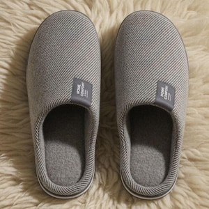 Pode incluir: Um par de chinelos cinzentos e listrados para uso interno. Cada chinelo tem um interior cinza e uma pequena etiqueta que diz "HOME COMFORT". Os chinelos estão em uma superfície texturizada de cor creme.