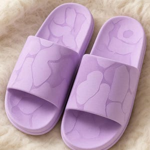May include: A pair of light purple slide sandals featuring a textured surface with a subtle, repeating organic camouflage-like pattern across the wide strap and the footbed. The sandals have a rounded toe design and a smooth, uniform matte finish.