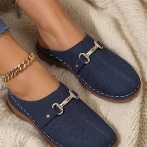 Può includere: Un paio di sabot blu navy con motivo floreale tagliato al laser sulla tomaia, un dettaglio metallico color argento a forma di morsetto e cuciture bianche a contrasto lungo il bordo della suola con suola piatta in gomma marrone.