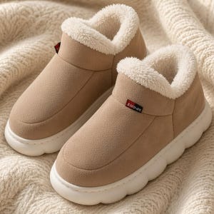 May include: A pair of tan slip-on slippers with a white faux-fur lining and a thick, white, rounded rubber sole. Each slipper features a small rectangular tag on the side with the word SPORT printed in white letters on a red and black background.