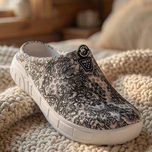 Può includere: Uno zoccolo senza lacci con un motivo floreale bianco e nero sulla tomaia. La scarpa presenta una spessa suola bianca e un emblema circolare nero sulla parte superiore con la parola NEXT stampata in bianco sopra un piccolo triangolo con puntini.
