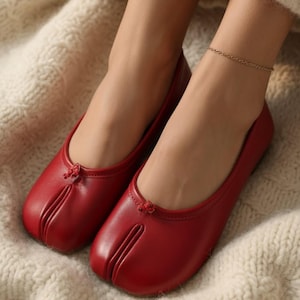 May include: A pair of deep red slip-on flats featuring a split-toe design at the front. Each shoe has a smooth, solid-colored exterior with a small, decorative knotted detail centered at the top of the opening, providing a clean, uniform look.