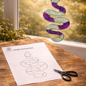 May include: A stained glass snake suncatcher in purple and green hangs near a window. A pattern for the suncatcher and scissors are on a wooden surface. Text at the bottom reads "INSTANT PDF/PNG DOWNLOAD."