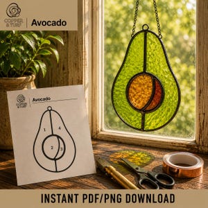 May include: A stained glass avocado suncatcher with green textured glass and a central orange pit, suspended by a metal chain. Beside it lies a printed pattern sheet labeled Avocado with numbered sections, a metal tool, and a roll of copper foil tape.