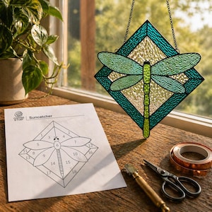 May include: A stained glass dragonfly suncatcher with green and teal accents hangs from a chain. A diagram labelled "Suncatcher" and craft tools, including scissors and copper tape, are on a wooden surface.