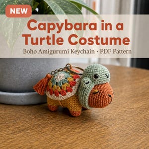 Op de afbeelding: Een gehaakte amigurumi-sleutelhanger van een bruine capibara met een saliegroene schildpaddenkap. De capibara heeft een kleurrijk granny square schild op de rug met een klein bijpassend kwastje. De tekst luidt: NEW Capybara in a Turtle Costume Boho Amigurumi Keychain PDF Pattern.