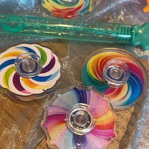 May include: A selection of rainbow-coloured fidget spinners with clear plastic centres. A green bubble wand and an orange frog-shaped toy are also visible. The spinners feature a spiral design in various colours.