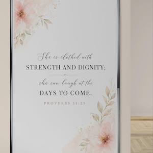 Könnte beinhalten: Gerahmter Kunstdruck mit schwarzem Rahmen. Der Druck zeigt den Text "She is clothed with STRENGTH AND DIGNITY; she can laugh at the DAYS TO COME. PROVERBS 31:25" in einer Schriftart. Aquarell-Blütenakzente in Rosa und Gold schmücken die Ecken.