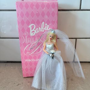Peut inclure: Poupée Barbie Millennial Porcelain, vêtue d'une robe de mariée blanche et argentée avec un voile, tenant un bouquet. La poupée est à côté d'une boîte rose avec le logo Barbie et le mot "Porcelain".