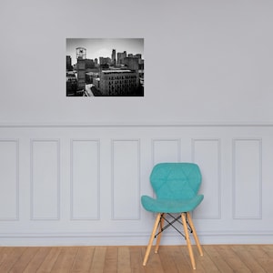 Pode incluir: Fotografia a preto e branco de uma paisagem urbana, com edifícios altos e uma torre do relógio. A imagem está montada numa parede acima de uma cadeira turquesa com pernas de madeira, criando um contraste.