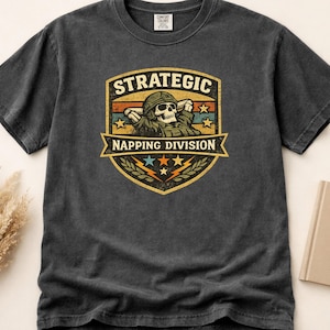 May include: Dark gray t-shirt with a distressed graphic. The graphic features a skull wearing a military helmet, with the words "STRATEGIC NAPPING DIVISION" in a shield design. The shirt has a crew neck and short sleeves.