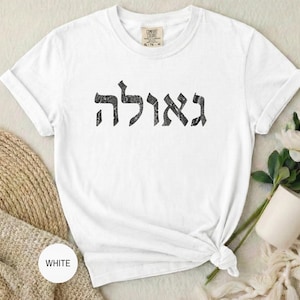 May include: White t-shirt with the Hebrew word "Ga'avah" printed in black. The shirt is knotted at the bottom and has a tag at the neckline. A white mug and flowers are in the background.