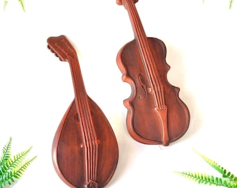 Pode incluir: Dois instrumentos musicais de madeira, um violino e um bandolim, são exibidos contra um fundo branco. O violino é marrom com um tom avermelhado e tem um design esculpido no corpo. O bandolim também é marrom com um tom avermelhado e tem um design esculpido no corpo. Ambos os instrumentos estão em foco e parecem ser feitos de madeira.