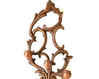 Pode incluir: Um castiçal de parede de madeira com um design decorativo e ornamentado. Possui três castiçais com um detalhe dourado na parte inferior.