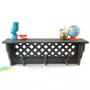 Op de afbeelding: Een donkerbruin houten plank met een traliewerkontwerp en drie haken. De plank toont een turquoise uilbeeldje, een kunstgebit, een gouden appel en een kleine wereldbol. De plank is een decoratieve opbergoplossing.