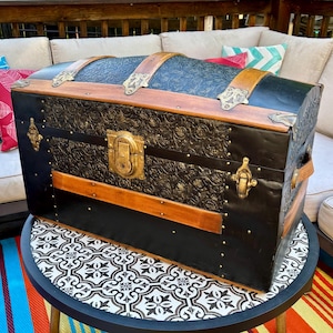 May include: An antique black trunk with ornate floral detailing and brass hardware. The trunk features wooden accents and leather straps. It sits on a small round table with a patterned top, placed on a colorful striped rug.