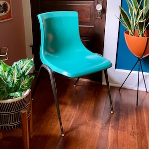 May include: A teal-colored vintage chair with a curved back and a seat. The chair has dark metal legs. The chair is in a room with hardwood floors and a dark wooden door. A potted plant is in the foreground.