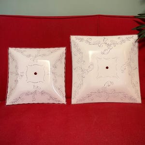 May include: Two square, white glass ceiling light fixtures with a decorative silver floral design. The fixtures are different sizes and have a small hole in the center. They are displayed on a red surface.
