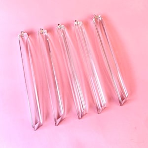 Può includere: Cinque prismi di cristallo trasparenti e rettangolari con un design sfaccettato, disposti diagonalmente su uno sfondo rosa. Ogni prisma ha un piccolo cappuccio di metallo nella parte superiore. I prismi sono lunghi circa 15 cm.