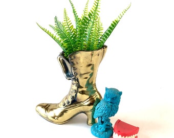 Pode incluir: Um vaso dourado em forma de bota com uma planta de samambaia verde dentro. Uma figura de coruja de cerâmica azul e um conjunto de dentes falsos de plástico vermelho estão sobre a superfície branca.