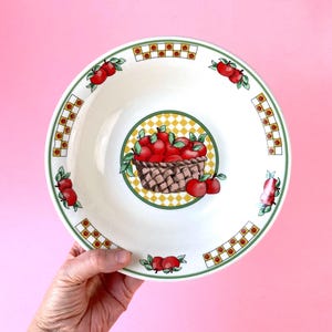 Può includere: Ciotola da portata in ceramica bianca con un motivo a mele. La ciotola presenta un bordo a scacchi verde e giallo, con illustrazioni di mele rosse sul bordo. Il centro della ciotola presenta un cestino di mele rosse.