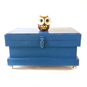 May include: A blue wooden storage chest with a latch and small wheels. A decorative owl figurine with large eyes sits on top. The chest is painted a solid blue color.