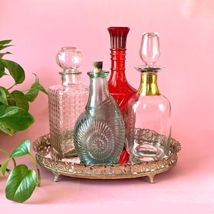 May include: An assortment of decorative glass bottles with stoppers, displayed on a silver-colored tray. The bottles vary in shape, color, and design, including clear, red, and blue-green hues. The tray is ornate with a floral pattern.