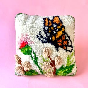 May include: A square, off-white, textured pillow with a butterfly design. The butterfly has orange, black, and white wings. The pillow also features pink, green, and tan floral accents. The edges are trimmed with green.