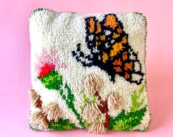 Pode incluir: Uma almofada quadrada e texturizada em branco sujo com um desenho de borboleta. A borboleta tem asas laranja, pretas e brancas. A almofada também apresenta detalhes florais rosa, verdes e bege. As bordas são aparadas em verde.