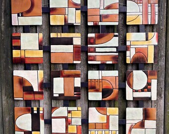 Pode incluir: Uma obra de arte de parede geométrica com dezasseis azulejos quadrados em tons de castanho, laranja, amarelo e branco. Os azulejos estão dispostos num padrão de grelha e montados numa moldura de metal preto. A obra é exibida contra uma cerca de madeira desgastada.