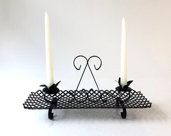Pode incluir: Castiçal de metal preto com duas velas brancas. O castiçal tem uma base retangular com um design de treliça e dois castiçais com detalhes florais.