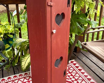 Pode incluir: Um armário alto e estreito de madeira vermelha com uma porta única que apresenta dois recortes em forma de coração cobertos por uma rede de arame. O armário tem um pequeno puxador redondo branco, dobradiças de metal preto e topo plano.