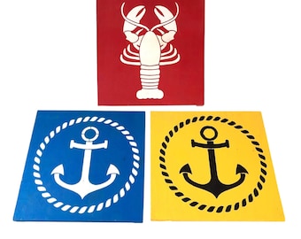 Pode incluir: Três placas quadradas de madeira com desenhos náuticos. Uma placa vermelha apresenta uma lagosta branca. Uma placa azul e uma placa amarela exibem cada uma uma âncora branca e preta, respectivamente, circundada por um desenho de corda branca e preta.
