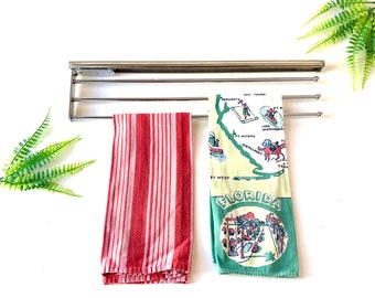 Pode incluir: Um suporte de toalha cromado com duas toalhas penduradas. A toalha à esquerda é listrada de vermelho e branco. A toalha à direita é verde e branca com um mapa da Flórida e o texto "FLORIDA" nela.