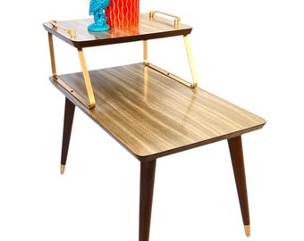 Pode incluir: Mesa lateral de dois níveis em estilo moderno de meados do século, com acabamento em grão de madeira escura. Possui detalhes em metal dourado e pernas com pontas douradas. Uma coruja turquesa e uma planta em vaso vermelha e verde estão no nível superior.