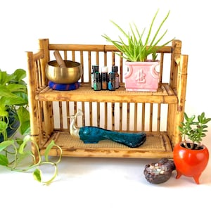 May include: A two-tiered bamboo shelf featuring a brass singing bowl on a blue cushion, several small essential oil bottles, and a pink ceramic planter with an aloe plant. A teal and gold peacock figurine sits on the bottom shelf.