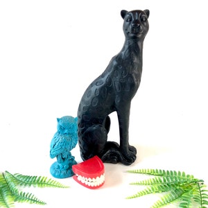 May include: A black panther figurine with a textured pattern sits next to a turquoise owl statue and a set of red dentures. Green fern leaves are arranged at the bottom. The objects are on a white background.