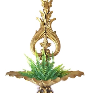May include: A gold-colored wall-mounted shelf featuring an ornate, elongated vertical backplate with scrolling leaf motifs and a scalloped, shell-shaped basin. A vibrant green artificial fern plant is placed inside the basin.