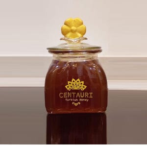 Puede incluir: Un tarro de cristal lleno de miel de color ámbar, coronado con una tapa decorativa amarilla en forma de flor. El tarro tiene una etiqueta dorada con el texto "CENTAURI Turkish Honey". El tarro está sobre una superficie reflectante.