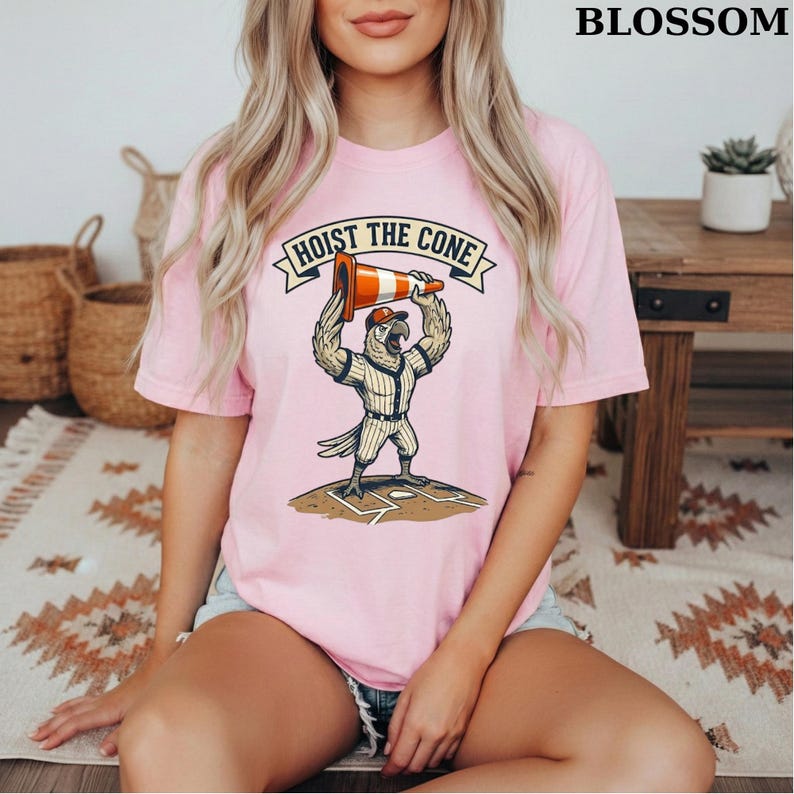 May include: Pink t-shirt with the text "HOIST THE CONE" above a cartoon parrot in a baseball uniform holding a traffic cone. The parrot is standing on a baseball field. The word "BLOSSOM" is printed at the top right.
