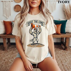May include: Ivory-colored t-shirt featuring a cartoon parrot in a baseball uniform holding a traffic cone above its head. The text "HOIST THE CONE" is printed above the parrot. The shirt is a casual, short-sleeved design.