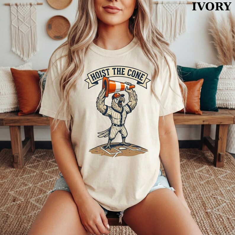 May include: Ivory-colored t-shirt featuring a cartoon parrot in a baseball uniform holding a traffic cone above its head. The text "HOIST THE CONE" is printed above the parrot. The shirt is a casual, short-sleeved design.