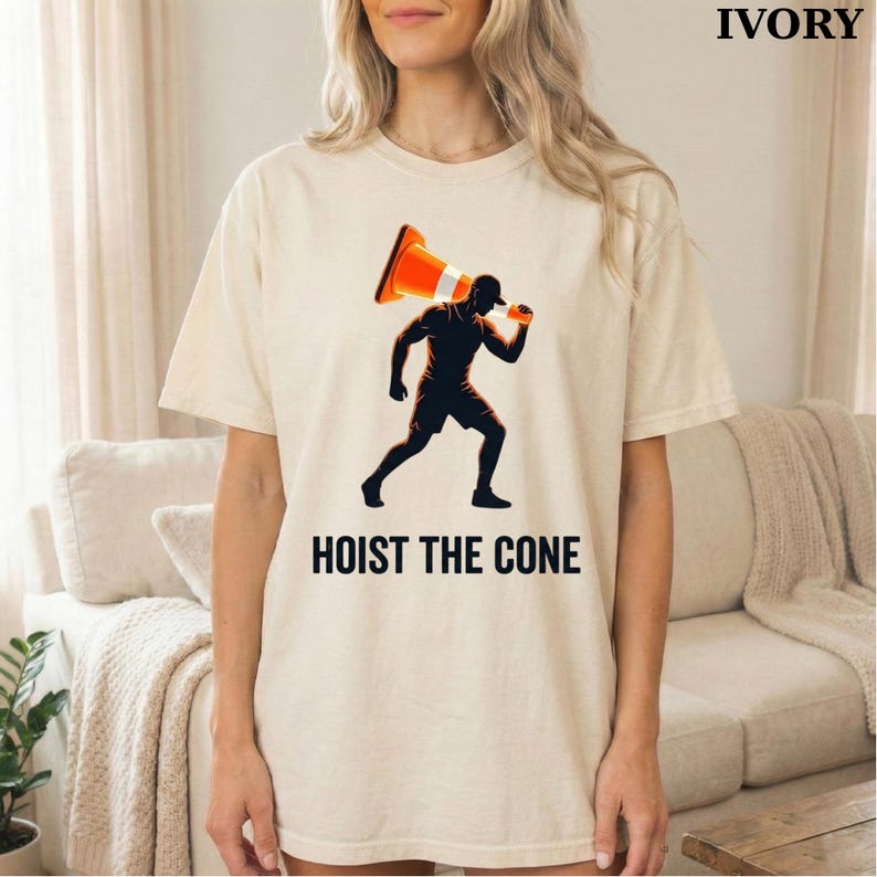 May include: Ivory-colored t-shirt featuring a graphic of a person carrying a traffic cone, with the text "HOIST THE CONE" in black. The graphic is in shades of orange and black. The shirt is a casual, short-sleeved style.