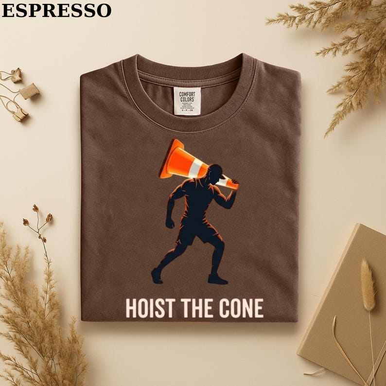 May include: Espresso-colored t-shirt with a graphic of a person carrying a traffic cone. The text "HOIST THE CONE" is printed in white. The shirt is made of Comfort Colors material.