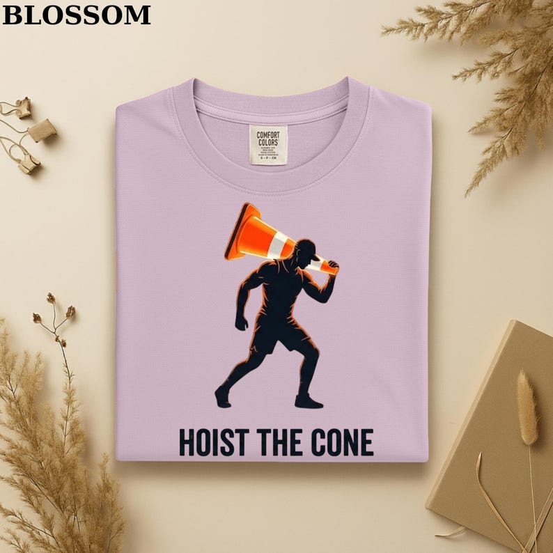 May include: Lilac Comfort Colors sweatshirt with a graphic of a person carrying a traffic cone. The person is in silhouette, and the cone is orange and white. The text "HOIST THE CONE" is printed below the graphic.
