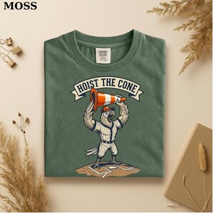 May include: Green t-shirt with a graphic of a baseball-playing bird lifting an orange and white traffic cone. The text "HOIST THE CONE" is above the bird. The shirt has a Comfort Colors tag in the upper right.